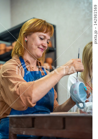 Woman over 50 painting handmade ceramic mug with brush in pottery workshop. Creative hobby represents active lifestyle and self expression in mature adulthood Woman over 50 painting handmade ceramic mug with brush in pottery workshop. Creative hobby represents active lifestyle and self expression in mature adulthood 142981569
