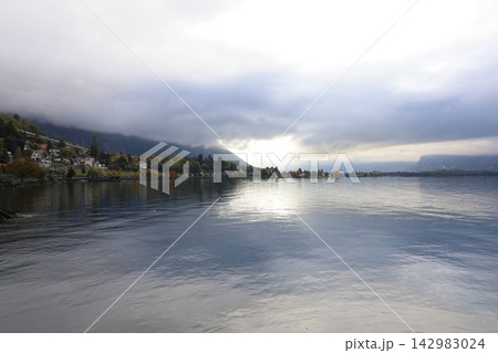 Moody autumn lake landscape with dramatic storm clouds, sunlight breaking through, misty mountain and lakeside village with colorful fall trees 142983024
