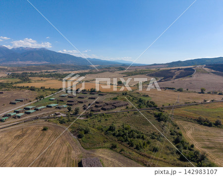 Heat power station site lies beside dry fields. Aerial view shows rural storage yards, roads, and power lines under clear sky, with mountains rising beyond industrial landscape and material storage. 142983107