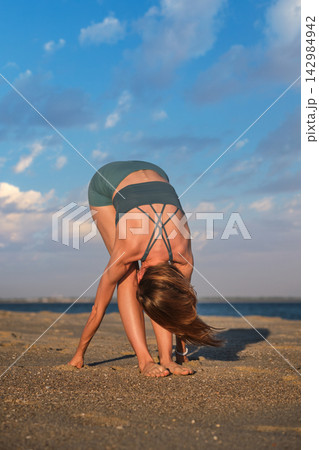Woman performs Uttanasana on a sandy beach at sunset near the ocean Woman performs Uttanasana on a sandy beach at sunset near the ocean 142984942