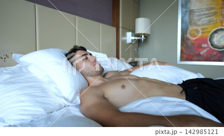 Young shirtless man watch cinema on TV lying on large bed in modern hotel room or home interior. Relaxed guy enjoying leisure, entertainment and comfortable lifestyle in comfortable place. Close up Young shirtless man watch cinema on TV lying on large bed in modern hotel room or home interior. Relaxed guy enjoying leisure, entertainment and comfortable lifestyle in comfortable place. Close up 142985121