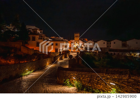 Illuminated church tower overlooks Ronda's rooftops from a cliffside stone bridge at night Illuminated church tower overlooks Ronda's rooftops from a cliffside stone bridge at night 142985900