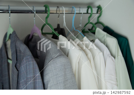 In the closet, checkered, dark and gray jackets hang on a chrome crossbar, close to a row of snow-white and light blouses on hangers. Concept: standard organization of clothes, contrast of textures In the closet, checkered, dark and gray jackets hang on a chrome crossbar, close to a row of snow-white and light blouses on hangers. Concept: standard organization of clothes, contrast of textures 142987592