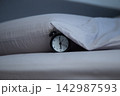 Close-up of a black alarm clock lying on a bed next to a pillow, partially covered by the edge of a gray blanket on a gray background. The concept of getting up in the morning, sleeping patterns, and 142987593