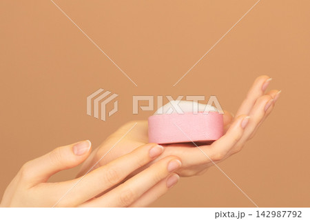 Close-up of female hands palm with pink solid face cleanser or hand foamy soap on isolated peach background. skin care advertising copy space. Close-up of female hands palm with pink solid face cleanser or hand foamy soap on isolated peach background. skin care advertising copy space. 142987792