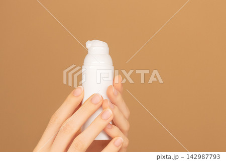 Female palms holding white bottle jar with face moisturizing cream dispenser on isolated peach background. Ready template for brand presentation. Skin care ad, copy space. Female palms holding white bottle jar with face moisturizing cream dispenser on isolated peach background. Ready template for brand presentation. Skin care ad, copy space. 142987793