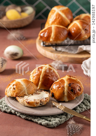 Vertical shot of glazed hot cross buns on a plate with one sliced and buttered in the foreground against a rustic kitchen backdrop with Easter eggs Vertical shot of glazed hot cross buns on a plate with one sliced and buttered in the foreground against a rustic kitchen backdrop with Easter eggs 142988197
