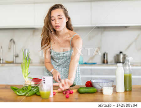 Young woman preparing salad and looking at smartphone in kitchen 142989204
