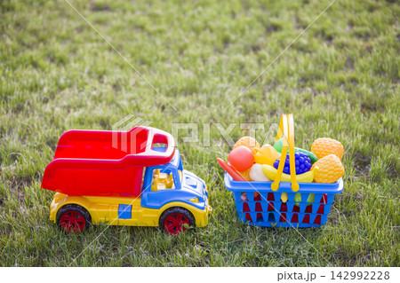 Car truckand a basket with toy fruit and vegetables. Bright plastic colorful toys for children outdoors on sunny summer day. Car truckand a basket with toy fruit and vegetables. Bright plastic colorful toys for children outdoors on sunny summer day. 142992228