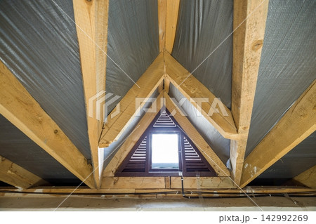 Attic of a building with wooden beams of a roof structure and a small window. Attic of a building with wooden beams of a roof structure and a small window. 142992269