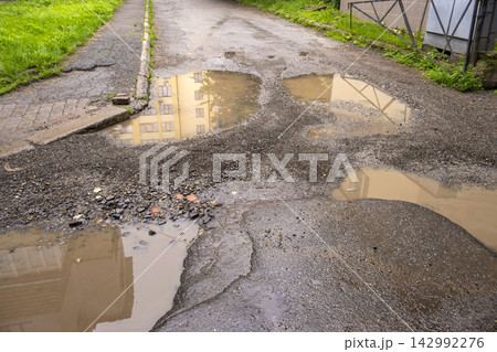 Large potholes filled with dirty rainwater on city street, creating a dangerous obstacle for cars while illustrating road decay and public infrastructure problems. 142992276