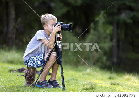 Young blond child boy sitting on tree stump on grassy clearing taking picture with tripod camera. Young blond child boy sitting on tree stump on grassy clearing taking picture with tripod camera. 142992778
