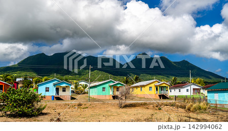 Row of vibrant colorful wooden houses in a rural Caribbean village with lush green mountains and cloudy blue sky in Saint Kitts and Nevis. Row of vibrant colorful wooden houses in a rural Caribbean village with lush green mountains and cloudy blue sky in Saint Kitts and Nevis. 142994062