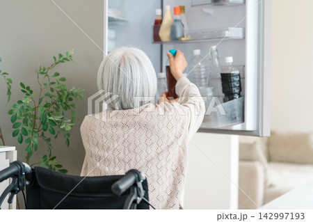 車椅子から冷蔵庫の飲み物を取り出すシニア女性。家事をこなす自立した高齢者の暮らし 車椅子から冷蔵庫の飲み物を取り出すシニア女性。家事をこなす自立した高齢者の暮らし 142997193