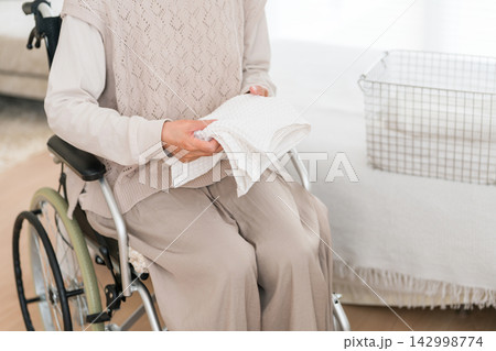 車椅子で洗濯物をたたむシニア女性。家事を続ける高齢者の自立生活と負担 車椅子で洗濯物をたたむシニア女性。家事を続ける高齢者の自立生活と負担 142998774
