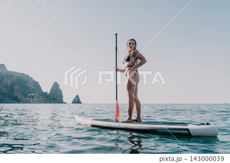 Woman Paddleboarding Ocean Mountains - A woman in a bikini stands on a paddleboard in the ocean with mountains in the background. Woman Paddleboarding Ocean Mountains - A woman in a bikini stands on a paddleboard in the ocean with mountains in the background. 143000039