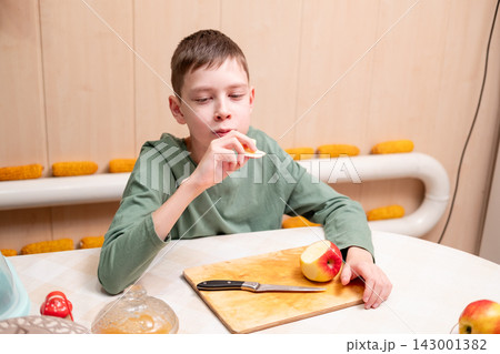 A young boy eating a slice of a red apple at a table with a wooden cutting board and a kitchen knife in front of him A young boy eating a slice of a red apple at a table with a wooden cutting board and a kitchen knife in front of him 143001382