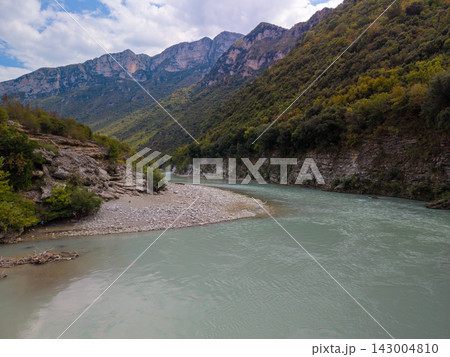 Vjosa River in Albania curves through rugged canyon terrain. Broad water, stony shoreline, and forested mountains create striking natural panorama with serene, remote atmosphere along gravel banks. Vjosa River in Albania curves through rugged canyon terrain. Broad water, stony shoreline, and forested mountains create striking natural panorama with serene, remote atmosphere along gravel banks. 143004810