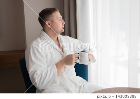 Young man in white bathrobe drinking coffee by window in hotel room Young man in white bathrobe drinking coffee by window in hotel room 143005888