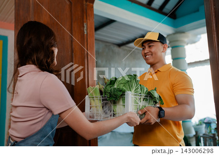 Smiling young man in orange shirt handing leafy greens crate to woman at home doorway, bringing fresh local food and community support Smiling young man in orange shirt handing leafy greens crate to woman at home doorway, bringing fresh local food and community support 143006298