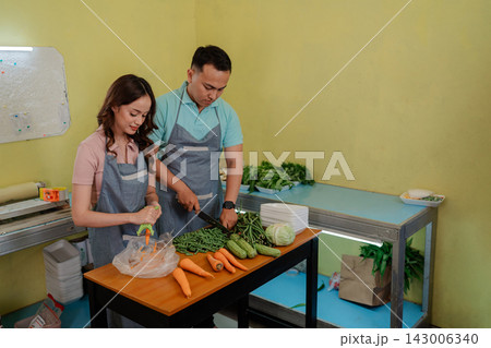 Two food workers preparing fresh vegetables for meal prep in small kitchen, focused teamwork, healthy cooking ingredients, aprons, cutting board, and simple workspace under soft indoor light Two food workers preparing fresh vegetables for meal prep in small kitchen, focused teamwork, healthy cooking ingredients, aprons, cutting board, and simple workspace under soft indoor light 143006340