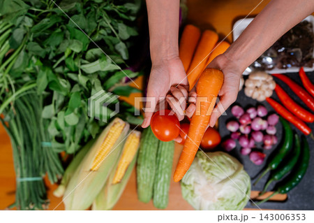 Hands holding carrot and tomato above colorful fresh vegetables on kitchen table with leafy greens, corn, cucumbers, onions, peppers, cabbage, garlic, and chili peppers Hands holding carrot and tomato above colorful fresh vegetables on kitchen table with leafy greens, corn, cucumbers, onions, peppers, cabbage, garlic, and chili peppers 143006353