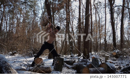 Shirtless young man chopping firewood in a sunny winter forest. Muscular guy working with axe in snowy woodland. Concept of strength, masculine, energy and harmony with nature during cold season 143007771