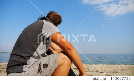 Happy couple hugs during sitting on wooden log on sandy beach. Romantic man kissing his beloved near coastline. Carefree lovers spending time together at resort. Concept of summer vacation or holiday 143007790