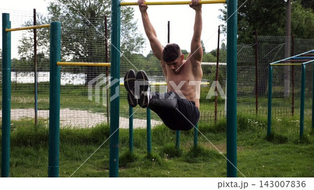 Athletic man trains abdominal muscles raising legs on horizontal bar at sports ground. Young sportsman exercising on pull-up bar outdoor. Sporty athlete working out at nature. Healthy lifestyle Athletic man trains abdominal muscles raising legs on horizontal bar at sports ground. Young sportsman exercising on pull-up bar outdoor. Sporty athlete working out at nature. Healthy lifestyle 143007836