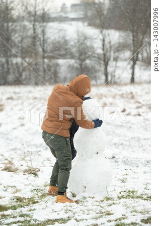 Building a snowman on a snowy day. A person in a brown jacket carefully packs snow onto the snowman's body. 143007996