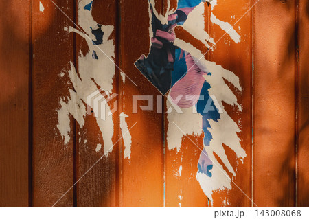 Torn Poster Remnants on Orange Surface. Layers of torn paper and old poster remnants peeling from weathered surface. Distressed urban texture with exposed fragments, scratches, irregular paper edges 143008068