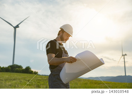 A man in a hard hat and overalls examines plans with wind turbines in the background during golden hour 143010548