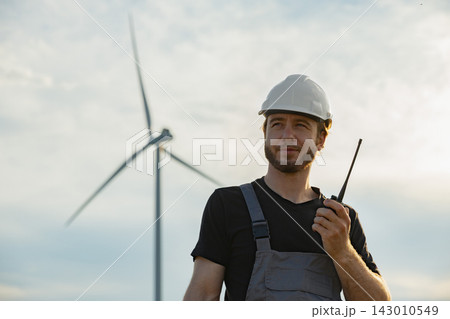 A Caucasian man in work overalls and a hard hat holds a radio while standing in front of a wind turbine on a cloudy day A Caucasian man in work overalls and a hard hat holds a radio while standing in front of a wind turbine on a cloudy day 143010549