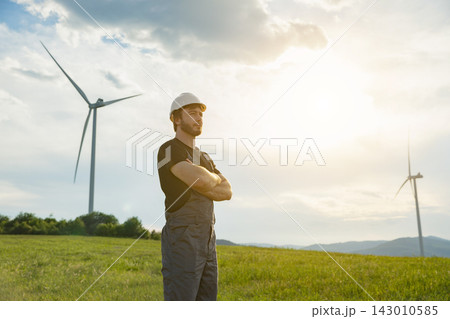 A Caucasian man wearing a hard hat and work overalls stands with his arms crossed in a grassy field with wind turbines in the background under a bright sky A Caucasian man wearing a hard hat and work overalls stands with his arms crossed in a grassy field with wind turbines in the background under a bright sky 143010585