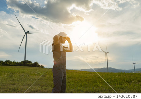 A man in work overalls and a hard hat stands in a grassy field with wind turbines in the background during a golden hour sunset A man in work overalls and a hard hat stands in a grassy field with wind turbines in the background during a golden hour sunset 143010587