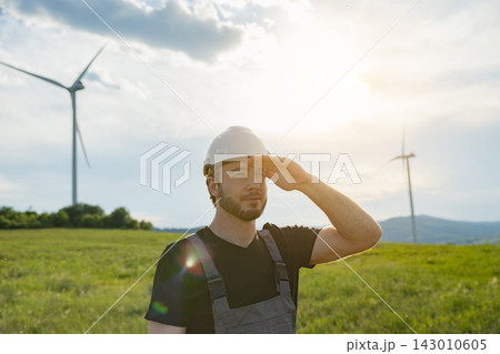 A Caucasian man wearing a hard hat and overalls stands in a green field with wind turbines generating renewable energy A Caucasian man wearing a hard hat and overalls stands in a green field with wind turbines generating renewable energy 143010605
