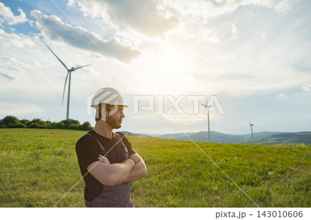 A Caucasian engineer wearing a hard hat and overalls stands with arms crossed in a grassy field overlooking wind turbines A Caucasian engineer wearing a hard hat and overalls stands with arms crossed in a grassy field overlooking wind turbines 143010606