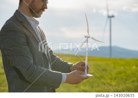 A Caucasian man in a suit jacket holds a miniature wind turbine model, symbolizing renewable energy development and planning A Caucasian man in a suit jacket holds a miniature wind turbine model, symbolizing renewable energy development and planning 143010635