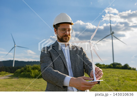 A Caucasian man wearing a hard hat and suit holds a miniature wind turbine model while standing in a field with large windmills behind him 143010636