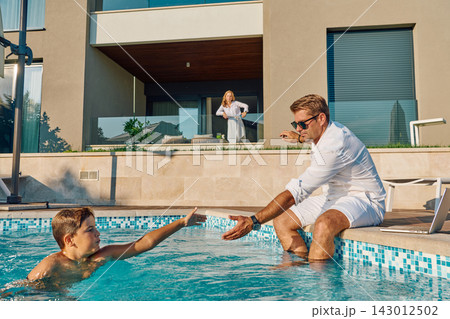 Family Enjoying Summer Pool Time at Modern Home While Father Helps Son and Mother Watches From Balcony Family Enjoying Summer Pool Time at Modern Home While Father Helps Son and Mother Watches From Balcony 143012502