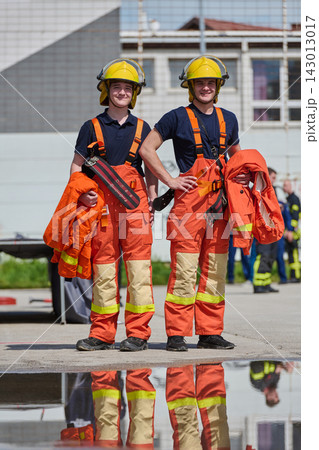 Two Young Firefighters In Orange Protective Uniforms And Helmets Standing Together Outdoors 143013017