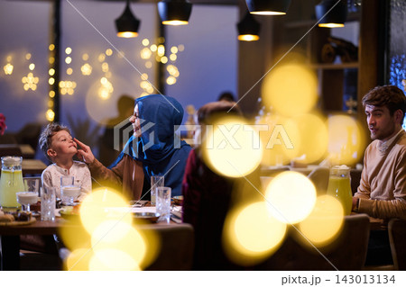 Muslim Family Sharing Warm Iftar Dinner With Child In Cozy Restaurant With Festive Lights Muslim Family Sharing Warm Iftar Dinner With Child In Cozy Restaurant With Festive Lights 143013134