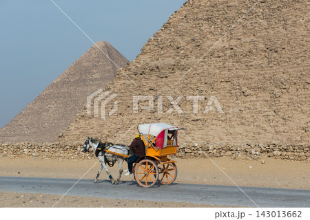 Authentic horse drawn carriage with orange wooden wheels and white canopy moving in profile past the towering limestone base wall of the Giza pyramids Authentic horse drawn carriage with orange wooden wheels and white canopy moving in profile past the towering limestone base wall of the Giza pyramids 143013662