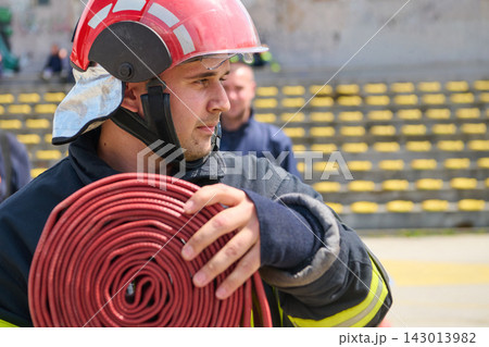 Firefighter Carrying Coiled Fire Hose Wearing Red Helmet And Protective Turnout Gear During Drill 143013982