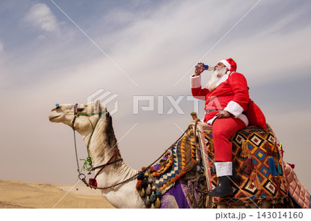 Santa Claus riding a colorfully decorated camel and drinking from a bottle in a vast sandy desert under overcast sky humorous Christmas holiday Santa Claus riding a colorfully decorated camel and drinking from a bottle in a vast sandy desert under overcast sky humorous Christmas holiday 143014160