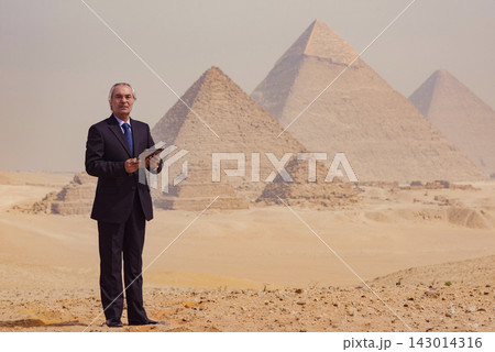 Composed silver haired senior executive in dark navy suit using digital tablet on sandy Giza plateau with iconic Great Pyramids in background ideal Composed silver haired senior executive in dark navy suit using digital tablet on sandy Giza plateau with iconic Great Pyramids in background ideal 143014316