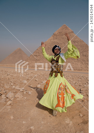 Tanoura dancer in vivid lime green and orange embroidered robe raises both arms overhead with skirt caught mid swirl against the Great Pyramids of 143014344