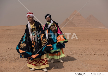 Folk costume portrait of two Egyptian male performers in vibrant embroidered robes and turbans smiling proudly before the iconic Giza Pyramids across 143014460