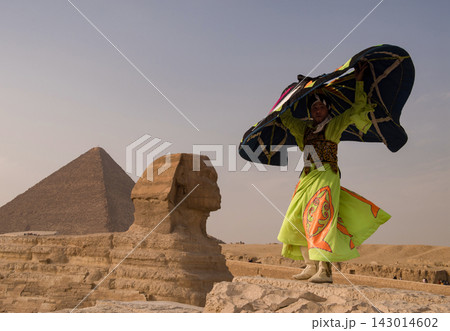 Joyful Egyptian folk performer in neon yellow embroidered costume dramatically raises a large kite on a stone terrace before the Great Sphinx and 143014602
