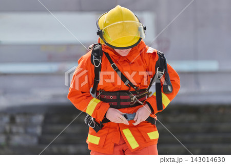 Firefighter In Orange Uniform And Yellow Helmet Fastening Safety Harness During Training Exercise Firefighter In Orange Uniform And Yellow Helmet Fastening Safety Harness During Training Exercise 143014630